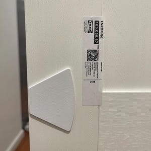 May include: Close-up of a white cabinet door with a textured surface. A white, triangular bumper is attached to the door. A label with the text "ENKÖPING" and the IKEA logo is affixed to the door.