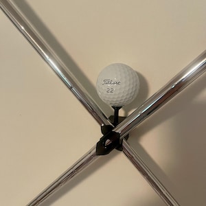May include: A white Titleist golf ball with the number 22 printed on it, held in place by a black metal holder. The holder is supported by four silver metal rods, creating a cross shape.