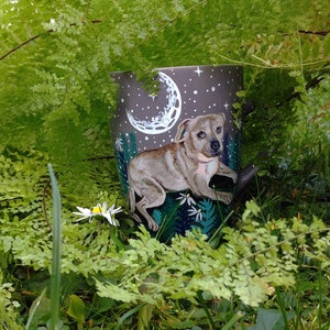 May include: A ceramic mug featuring a detailed illustration of a dog under a night sky with a crescent moon and stars. The mug has a dark gray background with white and green accents. The mug is surrounded by green foliage.