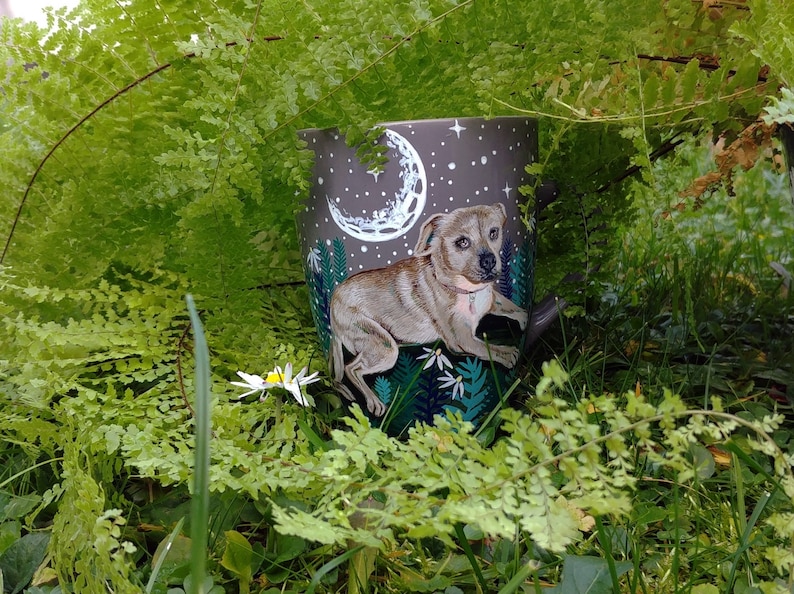 May include: A ceramic mug featuring a detailed illustration of a dog under a night sky with a crescent moon and stars. The mug has a dark gray background with white and green accents. The mug is surrounded by green foliage.