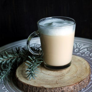 May include: A clear glass mug filled with a creamy, light brown beverage, topped with a thick layer of white foam. The mug sits on a wooden slice, placed on a decorative silver tray with sprigs of greenery.
