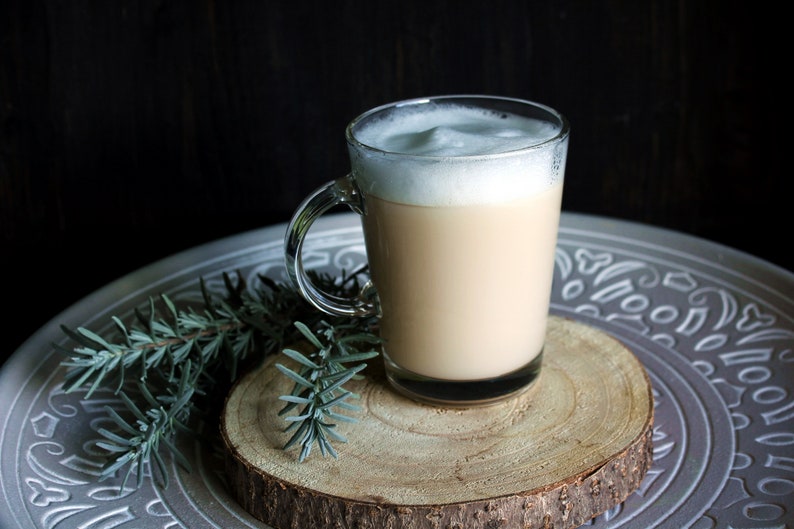 May include: A clear glass mug filled with a creamy, light brown beverage, topped with a thick layer of white foam. The mug sits on a wooden slice, placed on a decorative silver tray with sprigs of greenery.