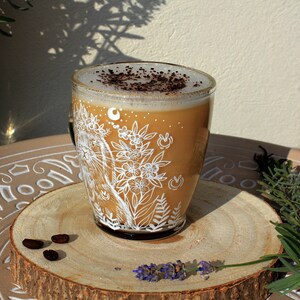 May include: A white ceramic mug with a floral design and a brown coffee drink inside. The mug is sitting on a wooden coaster with coffee beans on it.