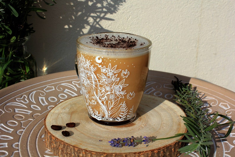May include: A white ceramic mug with a floral design and a brown coffee drink inside. The mug is sitting on a wooden coaster with coffee beans on it.