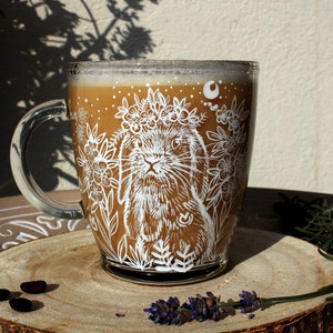 May include: A glass mug with a white design of a rabbit wearing a flower crown and surrounded by flowers. The mug is filled with a brown liquid and is sitting on a wooden surface.