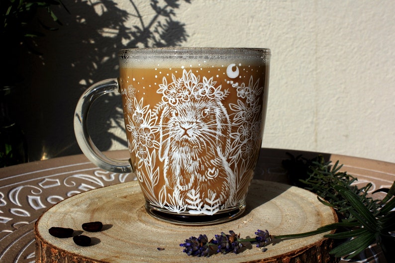 May include: A glass mug with a white design of a rabbit wearing a flower crown and surrounded by flowers. The mug is filled with a brown liquid and is sitting on a wooden surface.