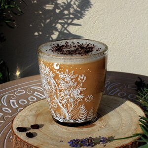 May include: A glass mug with a white floral design and a brown coffee drink topped with cocoa powder. The mug is sitting on a wooden coaster with coffee beans and lavender.
