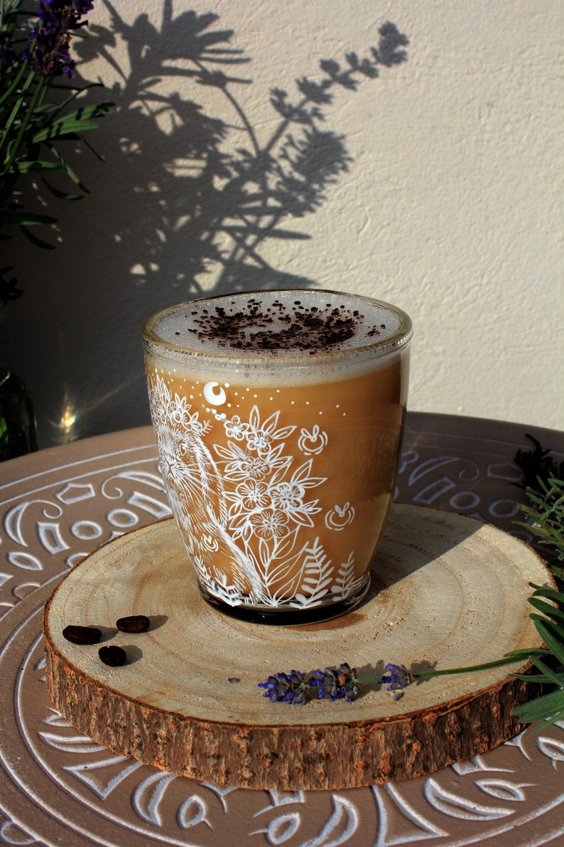 May include: A glass mug with a white floral design and a brown coffee drink topped with cocoa powder. The mug is sitting on a wooden coaster with coffee beans and lavender.