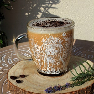 May include: A glass mug with a white bunny illustration and floral design. The mug is filled with a light brown beverage topped with a sprinkle of dark brown cocoa powder. The mug is sitting on a wooden coaster with a few coffee beans.