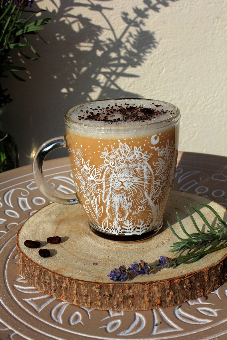 May include: A glass mug with a white bunny illustration and floral design. The mug is filled with a light brown beverage topped with a sprinkle of dark brown cocoa powder. The mug is sitting on a wooden coaster with a few coffee beans.