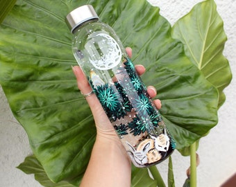 Puede incluir: Una botella de agua de vidrio transparente con una tapa plateada y un diseño pintado a mano. El diseño presenta una luna creciente, estrellas, follaje verde y dos pequeños animales durmiendo.