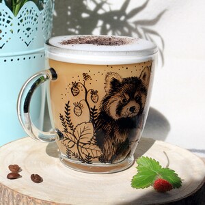 May include: A clear glass mug filled with a cappuccino, featuring a detailed illustration of a red panda and strawberry vines. The mug sits on a wooden coaster, with coffee beans and a strawberry nearby.
