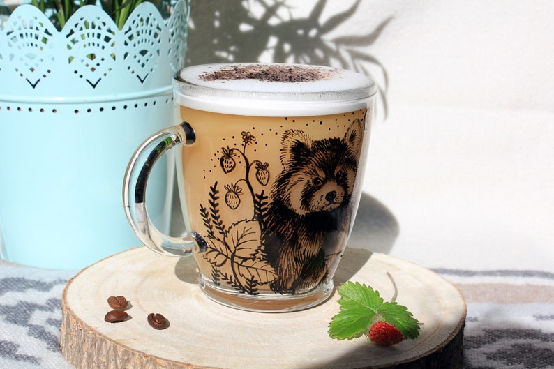 May include: A clear glass mug filled with a cappuccino, featuring a detailed illustration of a red panda and strawberry vines. The mug sits on a wooden coaster, with coffee beans and a strawberry nearby.
