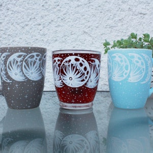 May include: Three ceramic mugs and a glass with a celestial design. The mugs are gray and light blue, and the glass is red. Each features a white illustration of moon phases and a speckled finish. A decorative plant is in the background.