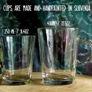May include: Two clear glass mugs with handles, one holding 8.4oz and the other 13.5oz. The mugs are on a wooden surface with a blurred, colorful background. Text at the top reads "CUPS ARE MADE AND HANDPAINTED IN SLOVENIA."
