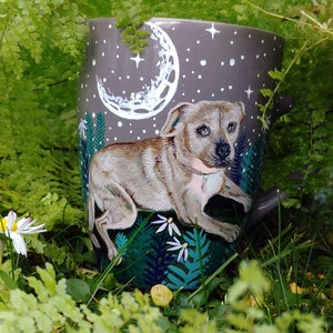 May include: A gray ceramic watering can featuring a painted dog design. The can has a night sky scene with a crescent moon and stars. The dog is light brown with a pink collar, and the can is surrounded by green foliage.