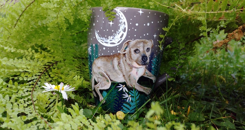 May include: A gray ceramic watering can featuring a painted dog design. The can has a night sky scene with a crescent moon and stars. The dog is light brown with a pink collar, and the can is surrounded by green foliage.