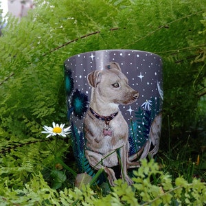 May include: A ceramic mug featuring a detailed illustration of a dog with a pink collar, set against a starry night sky background. The mug is surrounded by lush green ferns and a single daisy.
