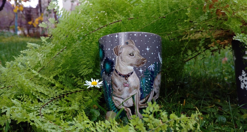 May include: A ceramic mug featuring a detailed illustration of a dog with a pink collar, set against a starry night sky background. The mug is surrounded by lush green ferns and a single daisy.
