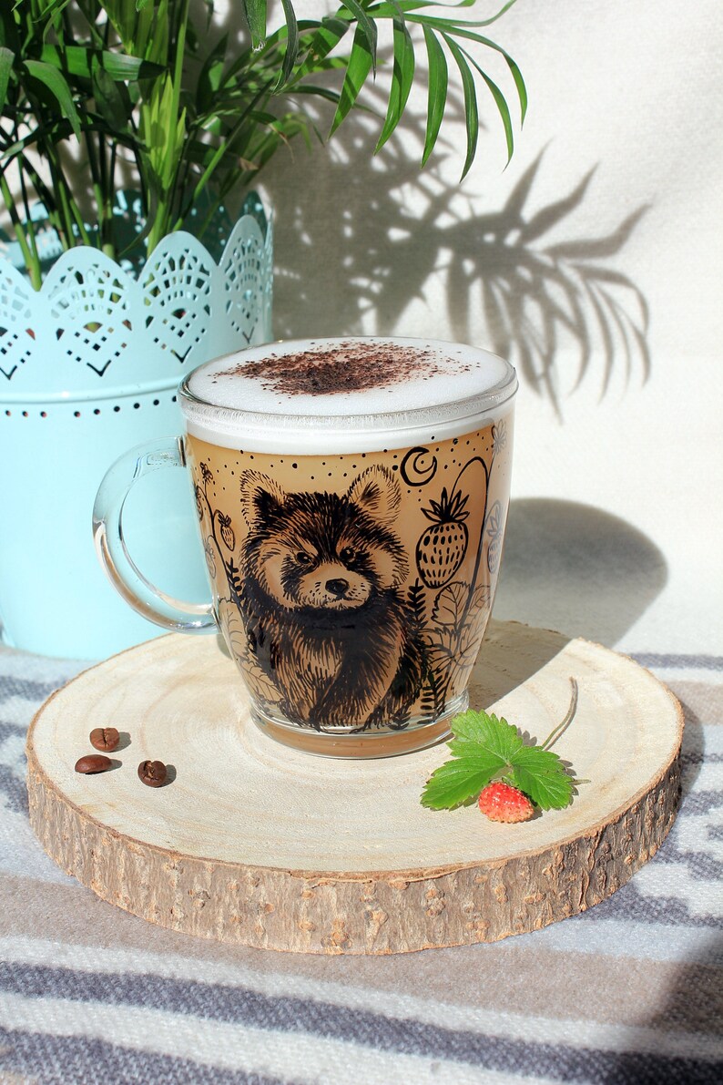 May include: A clear glass coffee mug with a red panda design, filled with a cappuccino. The mug sits on a wooden coaster with coffee beans and a strawberry. A light blue planter and green plants are in the background.