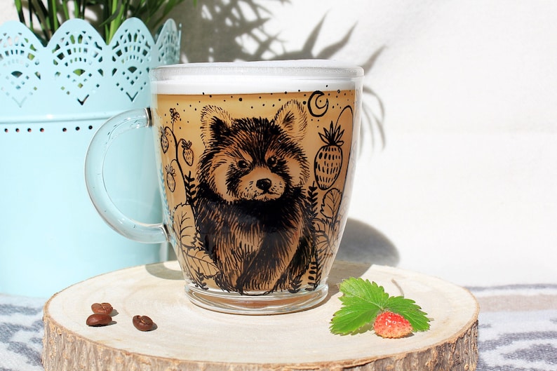May include: A clear glass mug with a handle, filled with a light brown beverage and foam. The mug features a detailed black illustration of a red panda surrounded by plants and fruit. The mug sits on a wooden slice.