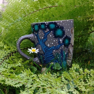 May include: A gray ceramic mug with a whimsical forest design featuring blue trees, teal foliage, and white stars. The mug is set amidst lush green ferns and grass, with a small white daisy in the foreground. The handle is gray.