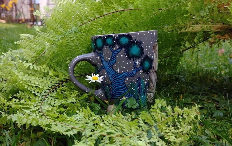 May include: A gray ceramic mug with a whimsical forest design featuring blue trees, teal foliage, and white stars. The mug is set amidst lush green ferns and grass, with a small white daisy in the foreground. The handle is gray.