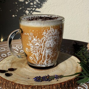 May include: A glass mug with a white design of a rabbit wearing a flower crown and surrounded by flowers. The mug is filled with a light brown beverage topped with a layer of foam and a sprinkle of cocoa powder. The mug is sitting on a wooden coaster with a few coffee beans scattered around it.