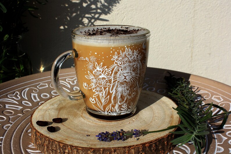 May include: A glass mug with a white design of a rabbit wearing a flower crown and surrounded by flowers. The mug is filled with a light brown beverage topped with a layer of foam and a sprinkle of cocoa powder. The mug is sitting on a wooden coaster with a few coffee beans scattered around it.