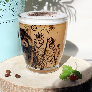 May include: A clear glass coffee mug with a cappuccino. The mug features a black illustration of a red panda and strawberry plants. The mug sits on a wooden coaster with coffee beans and a strawberry.