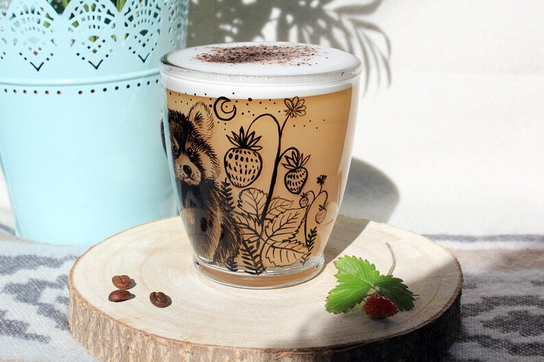 May include: A clear glass coffee mug with a cappuccino. The mug features a black illustration of a red panda and strawberry plants. The mug sits on a wooden coaster with coffee beans and a strawberry.