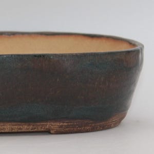 May include: A handmade, oval ceramic bonsai pot. The exterior is dark brown with teal hints and a lighter brown interior. The pot has a rustic, natural appearance, ideal for showcasing small plants.
