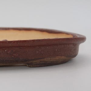 Op de afbeelding: Een bruine, ovale bonsai pot met een getextureerde buitenkant en een lichtbeige binnenkant. De pot heeft een laag profiel en is ontworpen voor het planten van kleine bomen of struiken. De rand is iets verhoogd en de basis heeft kleine voetjes.