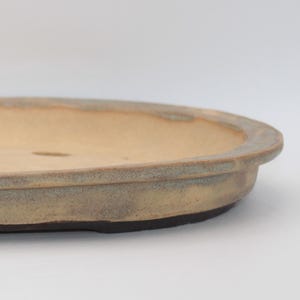 May include: An oval-shaped ceramic bonsai pot with a shallow basin and a wide, flat rim. The exterior features a muted, earthy beige and grey glaze, while the interior base is light tan and contains several drainage holes for plant care.