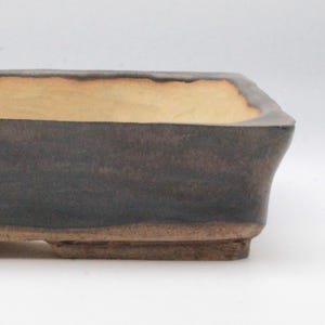 Könnte beinhalten: Ein rechteckiger Bonsai-Topf mit dunkelbrauner Außenseite und hellbraunem Innenraum. Der Topf hat eine leicht ausgestellte Form und steht auf kleinen rechteckigen Füßen. Die Kanten haben ein strukturiertes Aussehen.