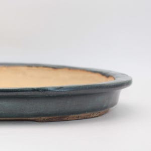 May include: A shallow, oval-shaped ceramic bonsai pot featuring a dark, glossy, deep blue-green glazed exterior rim and a contrasting unglazed, light beige interior base. The vessel has a low profile with a smooth, rounded edge and a flat bottom.