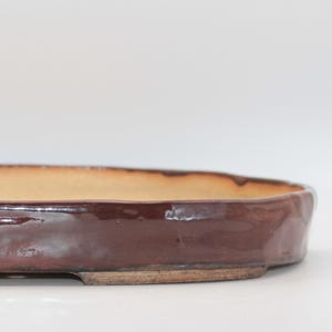 Könnte beinhalten: Ein glasierter, ovaler Bonsai-Topf. Die Außenseite ist ein tiefes Rotbraun, mit einem helleren, hellbraunen Innenraum. Der Topf hat ein niedriges Profil und ist mit einem flachen Boden und kleinen Füßen versehen. Das Gesamtdesign ist einfach und funktional.