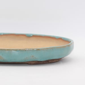 May include: An oval ceramic tray with a light turquoise glazed rim and a smooth, unglazed tan interior surface. The exterior sides feature a textured, rustic finish with visible variations in the blue glaze, resting on a dark brown base.