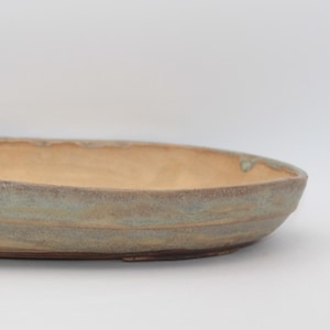 May include: An oval ceramic tray with a shallow basin and a textured, uneven rim. The exterior features horizontal bands of muted sage green and earthy tan tones, while the interior has a smooth, light beige finish with subtle green accents.