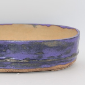 May include: An oval ceramic bonsai pot with a low profile and a wide opening. The exterior features a vibrant purple glaze with dark, mottled streaks and a horizontal ridge, while the interior and base reveal a natural, unglazed tan clay.