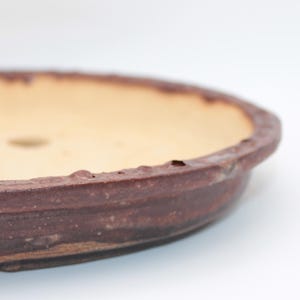 May include: An oval-shaped ceramic bonsai pot with a textured, dark brown exterior rim and a smooth, light beige interior basin. The shallow vessel features two drainage holes visible on the bottom surface, showcasing a rustic, earthy finish.