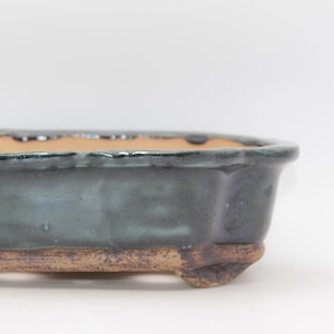 Könnte beinhalten: Ein ovaler Bonsai-Topf aus Keramik mit einer dunkel glänzenden, blau-grünen Glasur und einem gewellten Rand. Der Boden besteht aus unglasierter, strukturierter brauner Tonerde und hat vier kleine, stabile Füße, die das Gefäß anheben.