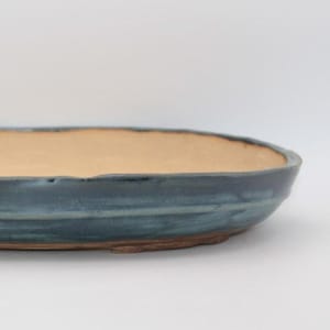 Peut inclure: Un pot à bonsaï ovale en céramique avec un bassin peu profond et un intérieur beige clair lisse. L'extérieur présente une glaçure bleue profonde et variée avec des stries horizontales et une base en argile sombre non émaillée.