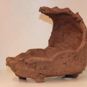 Op de afbeelding: Een bruine keramische plantenbak met een unieke, asymmetrische vorm. De plantenbak heeft een brede, open bovenkant en een gebogen, afgeronde basis. Hij is gemaakt van ruwe, getextureerde klei.