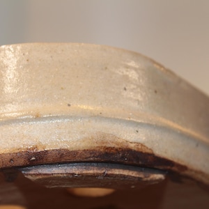 Op de afbeelding: Close-up van de bovenrand van een keramische bonsai pot. De pot is licht beige van kleur met een bruine rand. De pot heeft een drainagegat in de bodem.