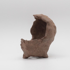 May include: A handmade, brown ceramic planter with a unique, organic shape. The planter has a rough, textured surface and a small, open interior. It stands on three small feet and is suitable for small plants or succulents.