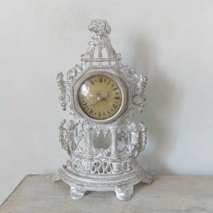 Puede incluir: Un reloj de chimenea ornamentado, de color blanquecino, con números romanos en la esfera. El reloj presenta figuras decorativas de querubines y volutas. El reloj mide aproximadamente 30 cm de altura y tiene una estética vintage.
