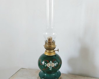 May include: A green glass oil lamp with a floral design and a clear glass chimney. The lamp has a brass base with a decorative design.