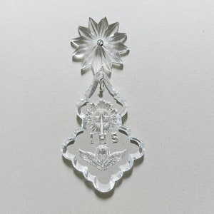 May include: A clear glass crystal ornament with a star-shaped top and a bell-shaped bottom. The bell has a cross and the letters "IHS" etched into it. The ornament also features a small angel with wings.