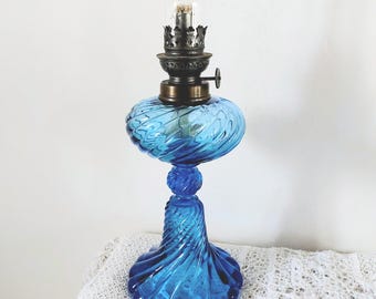 May include: A vintage oil lamp featuring a vibrant blue glass base and font with a distinct swirling ribbed pattern. The lamp includes a metallic burner assembly with a small adjustment knob and a clear glass chimney extending from the top.